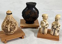 Three miniature Chinese decorative items grouped: cinnabar snuff bottle, metal vase, two Netsuke figurines, all on wooden stands.