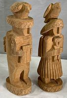 Two light-colored wooden statues side by side, showing full front and side profiles. Both stand on round carved bases and hold objects, with detailed geometric patterns carved into hats and clothing.