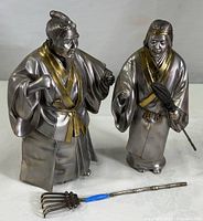 Full view of both sculptures together, showing one holding a rake detached and marked with blue tape for repair