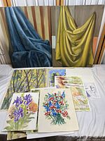 Photo showing two large unframed oil paintings featuring blue and yellow draped fabric, with various smaller watercolor paintings on paper arranged below them.