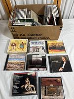 Photo of cardboard box containing multiple classical and Broadway audio CDs in jewel cases, showing front row of individual CDs displayed, including 'The Secret Garden', 'Mamma Mia!', Columbia Radio Symphony Orchestra, and more.
