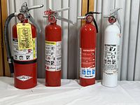 Front view of four fire extinguishers on a white surface, three red and one white with visible instruction labels and handles.