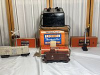 Full lot view with Lionel Trainmaster transformer box, transformer unit on top, Signal box and two rectangular Lionel Trains boxes, the brown caboose in front and a white train car to the left