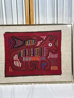 Full view of the framed red textile featuring a large animal with a bird on its back, decorated with multi-color striped embroidery.