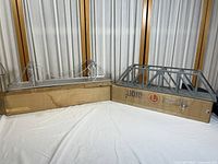 Pair of Lionel metal train bridges on original cardboard boxes, suspension and truss bridge style, placed on white cloth background with curtain behind