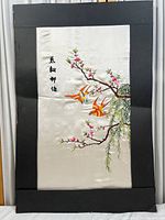 Silk embroidery piece mounted on black backing board showing two orange and yellow birds, blossoming branches with pink flowers and Chinese characters. Cream colored silk background visible with creases.