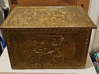 Front and top side view of antique brass embossed fireplace log box showing figures and some tarnished areas.