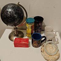 Photo showing black metallic globe, vintage Wilson tennis ball tin, Harry Potter mug, Deadpool cup, two animal mugs, glass ashtray and hair pin box on white table.