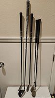 Five assorted golf clubs standing against wall, grips and shafts visible