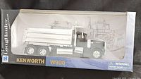 Model truck in original packaging showing name and brand. Black and silver Kenworth W900 Longhauler die-cast truck with dump bed visible through clear box front.
