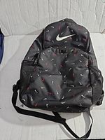 Front view of black backpack showing all-over Nike swoosh logos in white and red with a large white swoosh on top.