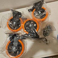 Four large casters in individual plastic bags with orange tires and black mounting plates, also includes a small bag with metal fasteners.