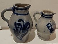 Two vintage German salt glazed pottery pitchers, showing gray body with cobalt blue floral and abstract designs, one larger and one smaller.