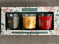 Boxed set of three Yankee Candle pillar candles labeled Peace, Love, Joy in festive holiday packaging with holly and berry design.