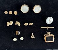 Top view of antique shirt studs, cufflinks, and pins arranged on black background showing varying sizes and materials including mother-of-pearl and metal.