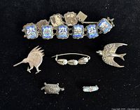 Top view of all jewelry pieces including an enamel decorated bracelet and five brooches on black background.