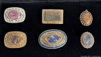Top view of six artisan made items including four belt buckles and two brooches or pendants, each with large polished stones in various colors and metal settings.