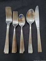 Five flatware pieces with dinner fork, salad fork, teaspoon, dinner spoon, and dinner knife laid flat on black background