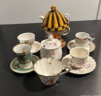Front view of assorted china teacups, saucers, a small teapot with floral design covered by a yellow and black knitted tea cozy, and a sugar bowl on a black surface.