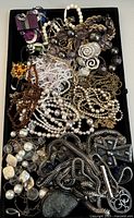 Overhead photo showing an assortment of costume jewelry, mostly necklaces with faux pearls, chains, and decorative pendants, in a black tray.