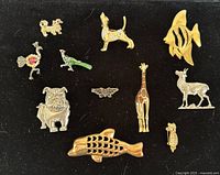 Top view of 12 animal-themed brooches arranged on black fabric including fish, dogs, birds, giraffe, deer, owl, and butterfly.