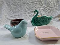 Four decorative ceramics displayed on white cloth: bluebird vase, green swan planter, small brown pot, pink plate with gold edge