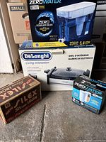Photo showing boxed DeLonghi indoor grill, ZeroWater water filter system with packaging, and vintage Silex steam iron box on floor.