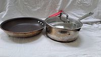Side-by-side view of the non-stick skillet and stainless steel saucepan with glass lid and red handle on a white surface.