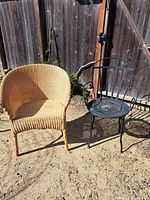 Side by side view of woven rattan chair and black metal chair outdoors on dirt surface with wooden fence background