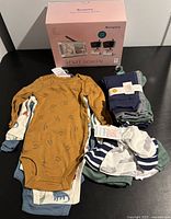 Moonybaby Split Screen baby monitor box and new baby clothes including onesies, pants, hats, socks, and mittens arranged on black surface