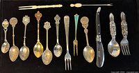 Photo showing full assortment of souvenir spoons, toddler bunny fork and spoon, small forks, and folding knife arranged on black background.