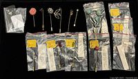 Stickpins displayed on black background, some loose and others in zip bags with labels, showing varied designs including floral, bird, gemstone themes