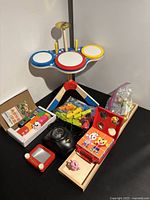 Overview photo showing all toys: wooden toy drum set with yellow drumsticks, puzzles including crocodile and box-fit, red Etch A Sketch, two Chinese block games, a black light projector, and a wooden pig figure.