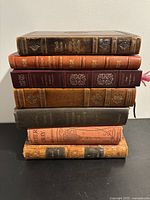 Stacked view of seven vintage books showing varied bindings including leather and cloth covers with embossed details.
