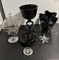 Front view of all six items arranged on black surface: clear covered compote bowl with floral etch, tall dark brown compote bowl, black and white ruffled vase, two crystal paperweights (star and oval), and black round candle holder.
