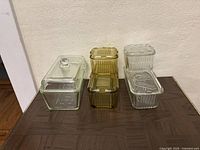 Side view of all glass containers and covers: clear rectangular loaf pan, amber ribbed containers stacked, clear vegetable pattern containers stacked.