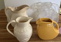 Photo showing all four pitchers arranged on wooden surface - large cream Crown Pottery pitcher, white Leedsware pitcher, yellow Fiesta pitcher, and clear glass swirl design pitcher.