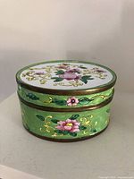 Front side view of the oval enamel trinket box showing floral decoration and brass trim.