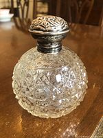 Front and side view of the hobnail cut glass perfume bottle with its ornate sterling silver lid attached, placed on a wooden surface.