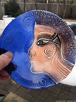 Blue crystal glass bowl featuring a painted profile of a face in blue and earthy tones, held against outdoor background.