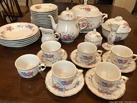 Full set of Hollohaza porcelain china displayed on wooden table, showing plates, teapot, creamer, sugar bowl, bonbonniere, candlestick, bell, and teacups with saucers.