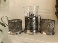 Three vintage Russian Soviet filigree tea glass holders displayed side by side, one holding a clear cut glass tea glass with embossed emblem on holder.