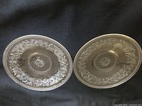 Two clear glass plates with finely engraved floral and bird designs, shown side by side on a dark background.