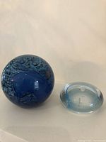 Two art glass paperweights shown together, one larger blue spherical with swirling patterns, one smaller clear blue bubble glass paperweight.