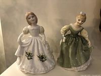 Front view of two Royal Doulton figurines, showing the April daisy figure in white dress and the Fair Maiden in green dress.