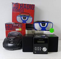 Photo showing Proscan Bluetooth CD micro system with dual speakers and three Proscan CD radio boomboxes, plus two boombox boxes, one red with image and text, another partially obscured.