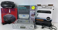 Photo showing the three items in the lot: the CD boombox on top of its box on the left, the Bluetooth under-counter CD clock radio in silver with its box in the center, and the black Sylvania HDMI upconvert DVD player with remote and cables on the right.