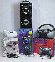 Photo of the three audio devices with their original boxes in background: black Bluetooth speaker, white CD boombox, black cassette player.