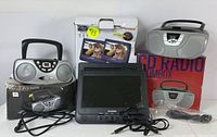 Image of two portable CD boomboxes, Sylvania dual-screen DVD player with power cords, remote controls, and original boxes.