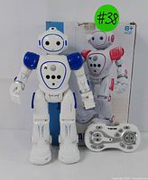 Front view of the white and blue interactive robot toy standing upright next to its remote controller and the original box showing a similar red robot model.